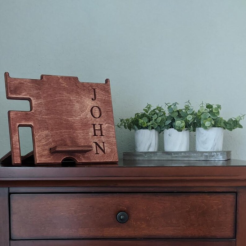 May include: A wooden phone stand with the name "JOHN" engraved on the side. The stand is sitting on a wooden table with a drawer and three small potted plants.