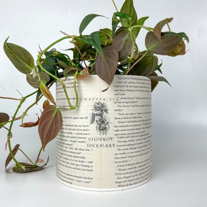 May include: A cylindrical plant pot with a book page design, filled with a lush green and burgundy plant. The pot features text and illustrations from a book, creating a unique decorative piece for home decor.