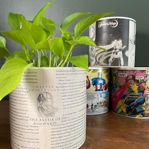 May include: A collection of decorative ceramic pots, each featuring unique designs. One pot displays text from "The Battle of Hogwarts," while others showcase comic book and anime art, including Captain America and Sailor Moon. The pots hold vibrant green plants.