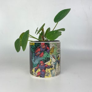May include: A cylindrical ceramic planter featuring a comic book design with characters in red, purple, and white. The planter holds a green plant with round leaves. The comic book design wraps around the planter.