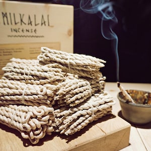Op de afbeelding: Stapels beige wierookstokjes, vastgebonden met touw, worden getoond op een houten oppervlak. Een houten doos met de tekst "SMILKALAI INCENSE" is zichtbaar. Rook stijgt op uit een kleine kom met een brandend wierookstokje.