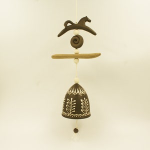 Puede incluir: Una campana de cerámica marrón con diseños de hojas blancas cuelga de una cuerda con cuentas de cerámica marrones y blancas. Un caballo de cerámica marrón y una espiral de cerámica marrón están unidos a la cuerda por encima de la campana.