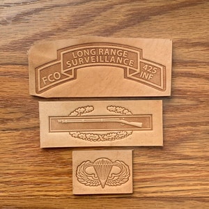 May include: Three leather patches with military insignia. The top patch is tan with the text "LONG RANGE SURVEILLANCE FCO 425 INF". The middle patch is tan with a rifle and laurel wreath. The bottom patch is tan with a parachute and wings.