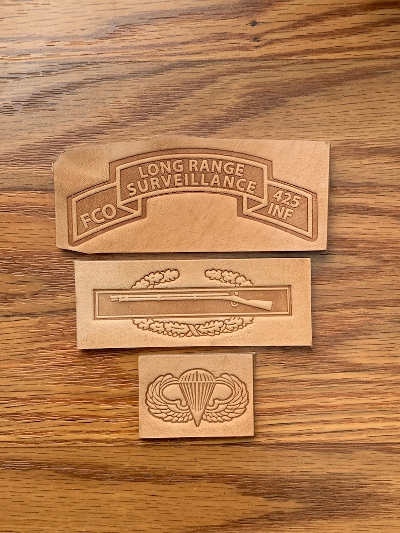 May include: Three leather patches with military insignia. The top patch is tan with the text "LONG RANGE SURVEILLANCE FCO 425 INF". The middle patch is tan with a rifle and laurel wreath. The bottom patch is tan with a parachute and wings.