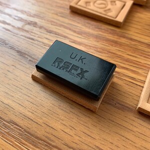 K&ouml;nnte beinhalten: Ein schwarzer rechteckiger Stempel mit dem Text "U.K. RSFX LASER PRODUCTS" auf einer braunen Lederoberfl&auml;che.
