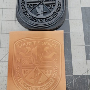 May include: A tan leather patch with the words "Sheriff's Office" in a curved banner at the top. The patch features a shield with a Spartan helmet, an American flag, and a goose. The words "Currituck County" are at the bottom of the patch, with the numbers "16" and "68" on either side of the goose.