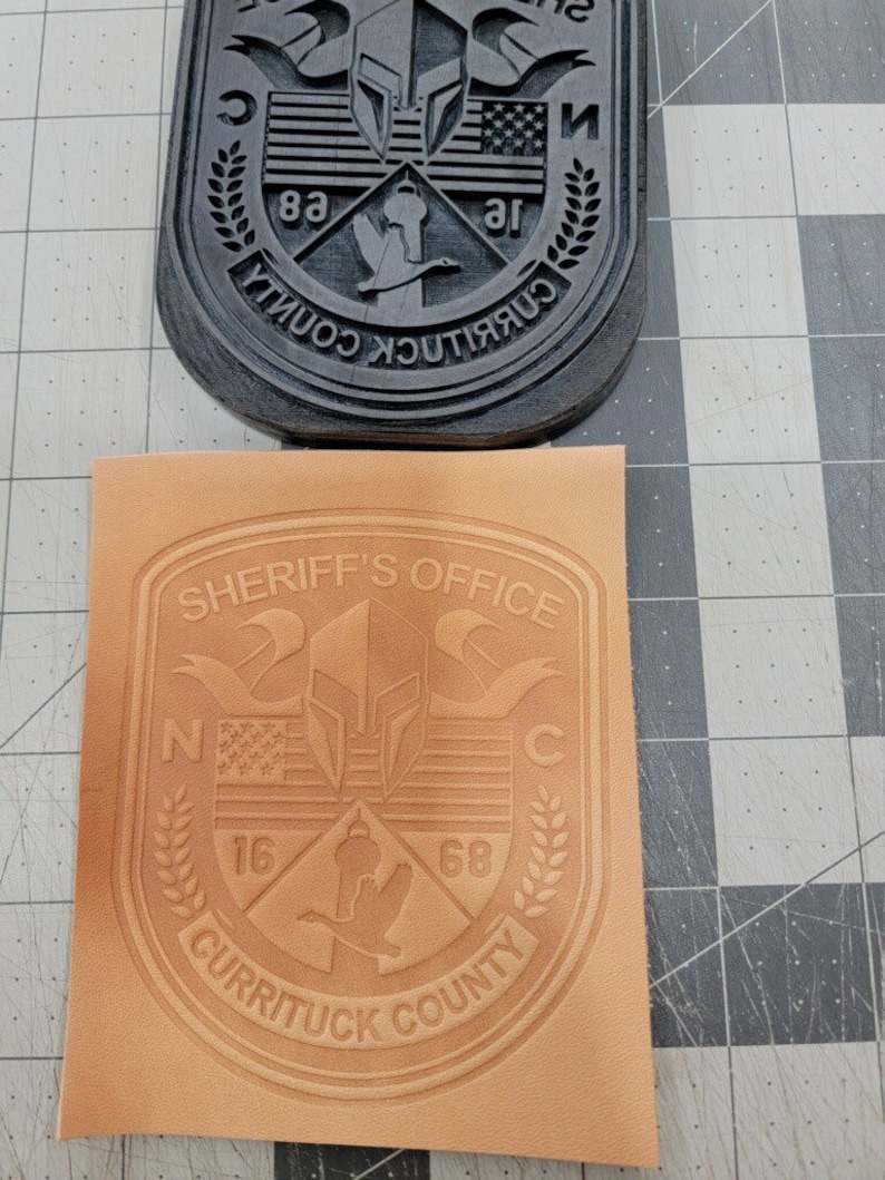 May include: A tan leather patch with the words "Sheriff's Office" in a curved banner at the top. The patch features a shield with a Spartan helmet, an American flag, and a goose. The words "Currituck County" are at the bottom of the patch, with the numbers "16" and "68" on either side of the goose.