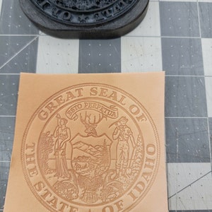May include: A tan leather stamp with the Great Seal of the State of Idaho. The seal features a woman holding a staff, a deer, a man holding a pickaxe, and mountains. The text "Great Seal of the State of Idaho" is around the outside of the seal.