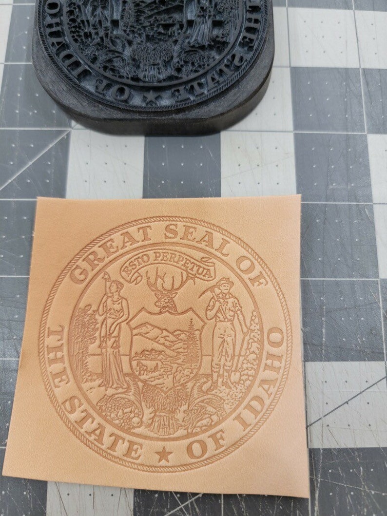 May include: A tan leather stamp with the Great Seal of the State of Idaho. The seal features a woman holding a staff, a deer, a man holding a pickaxe, and mountains. The text "Great Seal of the State of Idaho" is around the outside of the seal.