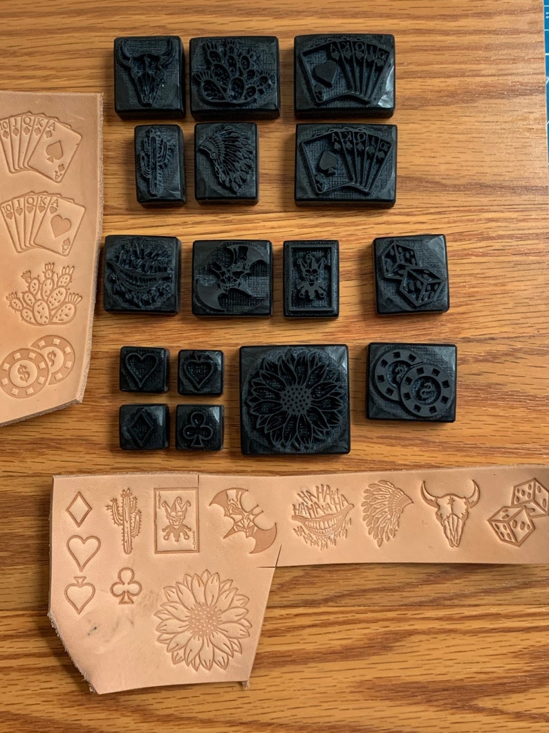 May include: A set of 20 black rubber stamps with various designs, including playing cards, cacti, skulls, dice, and a sunflower. The stamps are arranged on a wooden surface with a piece of leather next to them.