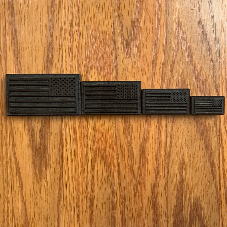 May include: Four rectangular black patches, each featuring a raised, molded design of the American flag. The patches are arranged in a horizontal row, decreasing in size from left to right, showcasing a consistent monochromatic aesthetic.