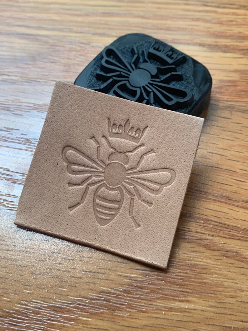 May include: A close-up of a leather stamp with a bee design. The bee is wearing a crown and has a striped body. The stamp is on a piece of brown leather.