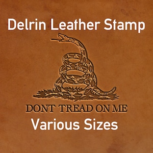 Könnte beinhalten: Delrin-Lederstempel mit einem Motiv einer aufgerollten Klapperschlange, der Text lautet "DONT TREAD ON ME" und "Various Sizes". Der Stempel ist auf einer braunen Lederoberfläche geprägt.