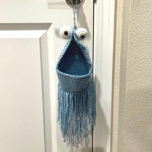 Könnte beinhalten: Ein blauer, gehäkelter Hängekorb mit Fransen unten und zwei weißen Glitz Augen. Der Korb hängt an einem Türknauf.