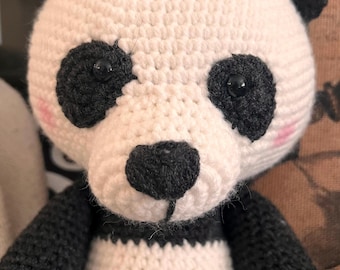 Puede incluir: Un oso panda hecho a mano con ganchillo, con cuerpo blanco, detalles negros e interior de las orejas rosa. El panda tiene ojos negros y nariz negra. Los brazos y las piernas del oso son negros y blancos. Es un peluche.