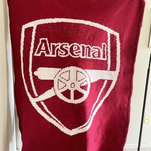 May include: A rectangular crochet blanket in deep red featuring the white Arsenal football club crest in the centre. The design includes the word Arsenal in white lettering above a white cannon graphic, all rendered in a consistent yarn texture.
