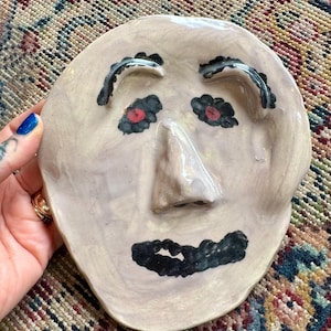 May include: A hand-sculpted ceramic face plate with a glossy light beige finish. The piece features a prominent raised nose, two black arched eyebrows, two black-rimmed eyes with red centers, and a wide black mouth painted on the surface.