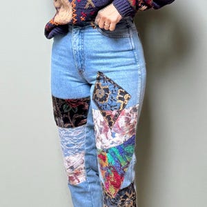 May include: Light blue denim jeans featuring various fabric patches sewn onto the front legs. The patches include floral, lace, and geometric patterns in black, white, red, green, and gold tones. The jeans have a high-waisted fit and cuffed hems.
