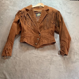 May include: A brown suede jacket featuring long fringe detailing across the chest and shoulders. The jacket has a notched collar, three front buttons, and lace-up details on the outer cuffs. It is displayed on a wooden hanger with a visible inner label.