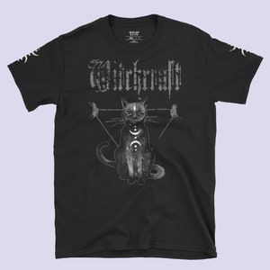 Pode incluir: Camiseta preta com um gráfico de um gato preto sentado em um triângulo com luas de cada lado. O texto "Witchcraft" está acima do gato.