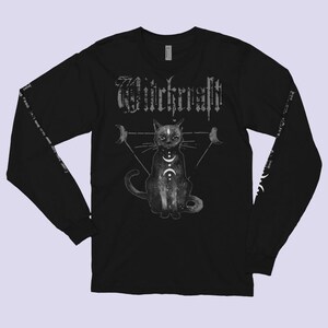Pode incluir: Camiseta preta de manga comprida com um gr&aacute;fico branco de um gato sentado em um tri&acirc;ngulo com luas e a palavra "Witchcraft" acima do gato.