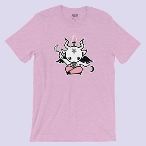 Pode incluir: Uma camiseta rosa com uma ilustração em preto e branco de um diabo de desenho animado com um pentagrama na testa, segurando uma vela e sentado em um coração. O texto "CRUEL AND  K.L.I.T.H" está impresso na etiqueta.