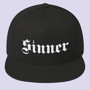 Puede incluir: Una gorra de b&eacute;isbol negra y blanca con un dise&ntilde;o bordado en blanco que dice "Sinner" en una fuente g&oacute;tica.