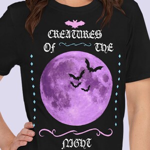 以下が含まれることがあります： 黒地に紫色の月とコウモリが描かれたTシャツ。ピンクと黒で「Creatures of the Night」の文字が書かれている。
