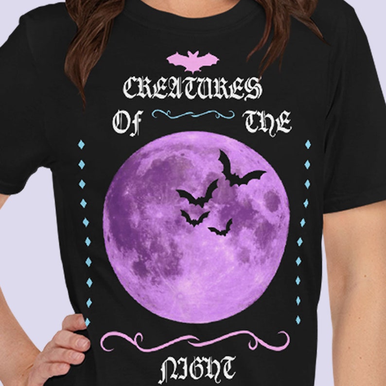 以下が含まれることがあります： 黒地に紫色の月とコウモリが描かれたTシャツ。ピンクと黒で「Creatures of the Night」の文字が書かれている。