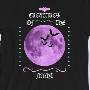 以下が含まれることがあります： 黒いTシャツに紫色の月とコウモリが描かれており、ピンク色の文字で「Creatures of the Night」と書かれています。