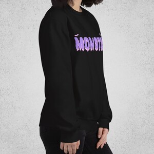 Pode incluir: Camisola de moletom preta com um gr&aacute;fico roxo de "MONSTER" com morcegos acima do texto.