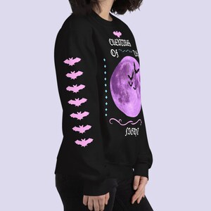 May include: Black sweatshirt with a purple moon and bats graphic. The text "Creatures of the Night" is printed in a gothic font. The sleeve has a row of pink bats.