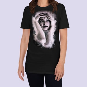 May include: Black t-shirt with a watercolor-style image of a woman with a cross on her forehead and a black mouth. The woman is covering her face with her hands.