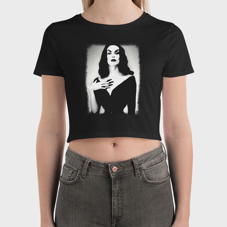 May include: Black cropped t-shirt with a white graphic of a woman in a black dress. The woman has dark hair and is looking directly at the viewer.