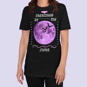 以下が含まれることがあります： 紫色の月とコウモリが描かれた黒いTシャツ。ピンクと白で「Creatures of the Night」の文字が書かれている。