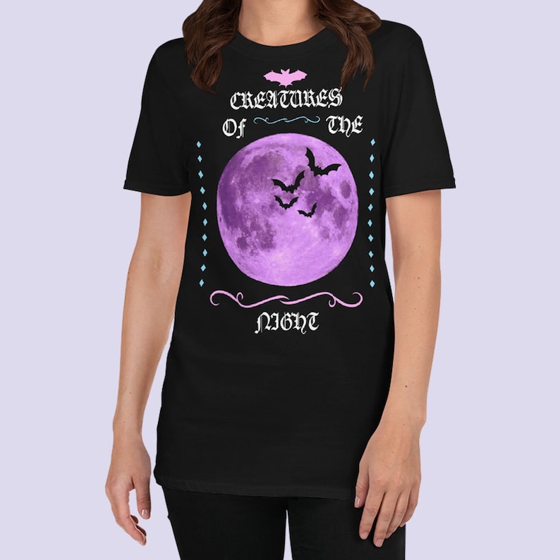 以下が含まれることがあります： 紫色の月とコウモリが描かれた黒いTシャツ。ピンクと白で「Creatures of the Night」の文字が書かれている。