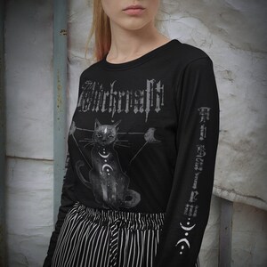 Pode incluir: Camiseta preta de manga comprida com uma estampa gr&aacute;fica de um gato preto com uma lua crescente e estrelas. O texto "Witchcraft" &eacute; impresso em uma fonte g&oacute;tica na frente da camiseta.