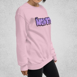 Pode incluir: Uma camisa de moletom rosa com um gr&aacute;fico roxo "MONST" estampado na frente.