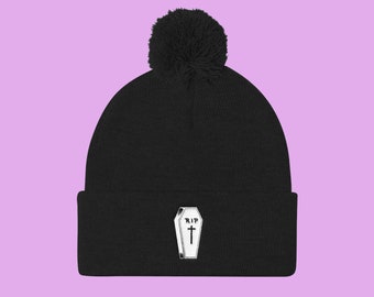Puede incluir: Gorro negro con un pompón esponjoso en la parte superior y un ataúd bordado en blanco con una cruz y el texto "RIP" en la parte delantera.
