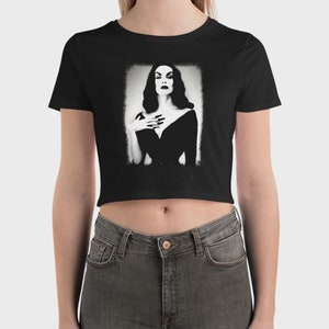 May include: Black cropped t-shirt with a white graphic of a woman in a black dress. The woman has dark hair and is looking directly at the viewer.
