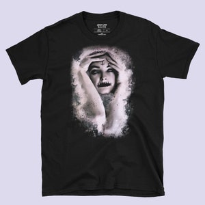 May include: Black t-shirt with a graphic print of a woman with a cross on her forehead and a painted mouth. The graphic is in a white and gray watercolor style.