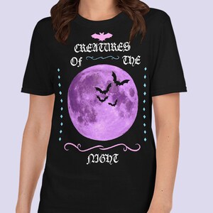 以下が含まれることがあります： 黒地に紫色の月とコウモリが描かれたTシャツ。ピンクと白で「Creatures of the Night」の文字が書かれている。