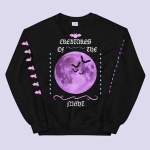 May include: Black sweatshirt with a purple moon and bats printed on the front. The text "Creatures of the Night" is printed in pink below the moon. The sleeves have a pink and white pattern.