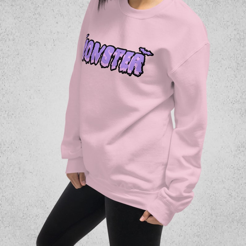 Pode incluir: Uma camisa de moletom rosa com um gr&aacute;fico roxo de "MONSTER" com um design de morcego.