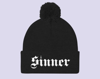 Puede incluir: Gorro negro con un pompón blanco y la palabra "Sinner" en letras góticas blancas.