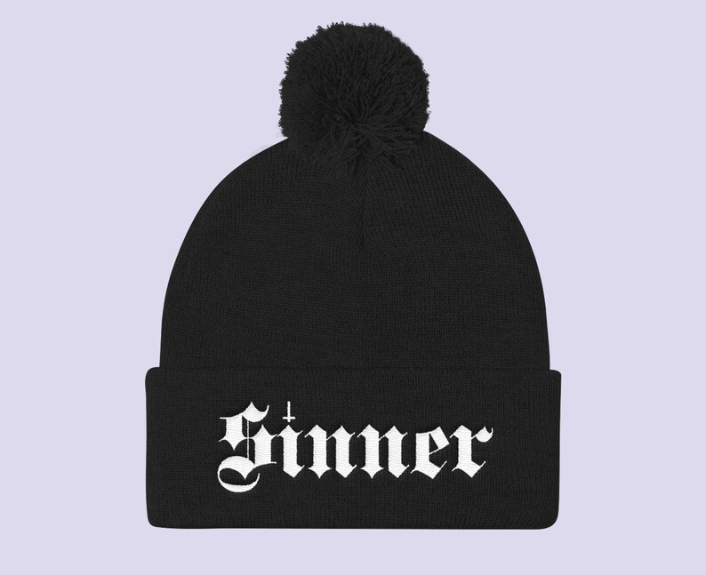 May include: Black beanie with a white pom pom and the word "Sinner" in white gothic lettering.