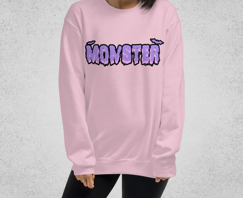 Pode incluir: Uma camisa de moletom rosa com a palavra "MONSTER" impressa em roxo com morcegos de cada lado da palavra.