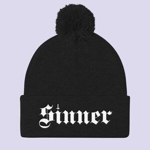 May include: Black beanie with a white pom pom and the word "Sinner" in white gothic lettering.
