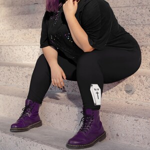 May include: Black leggings with a white coffin graphic that says "RIP" on the left thigh. The leggings are worn by a person wearing purple boots.