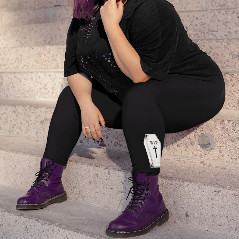 May include: Black leggings with a white coffin graphic that says "RIP" on the left thigh. The leggings are worn by a person wearing purple boots.
